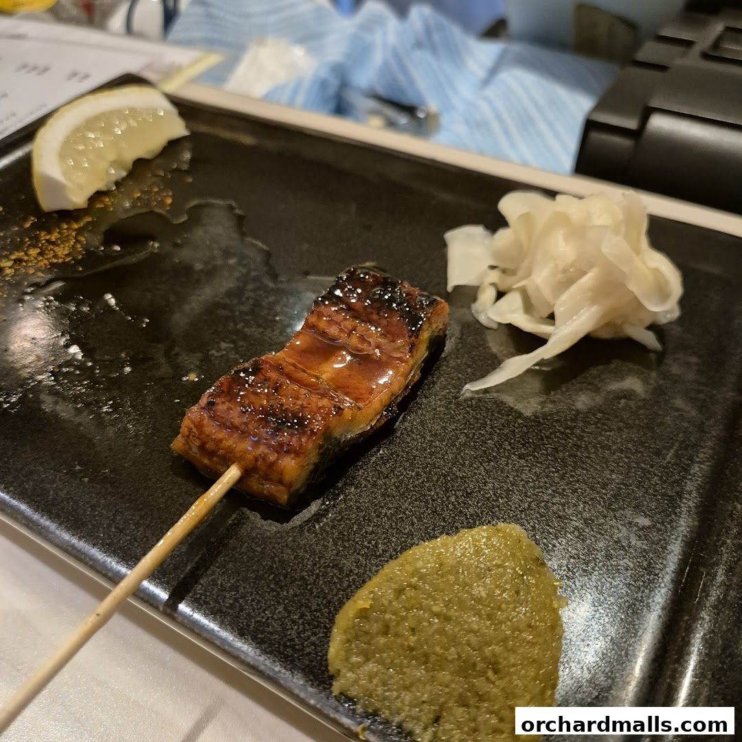 A glazed yakitori skewer with lemon, pickled ginger, and wasabi on a black plate.