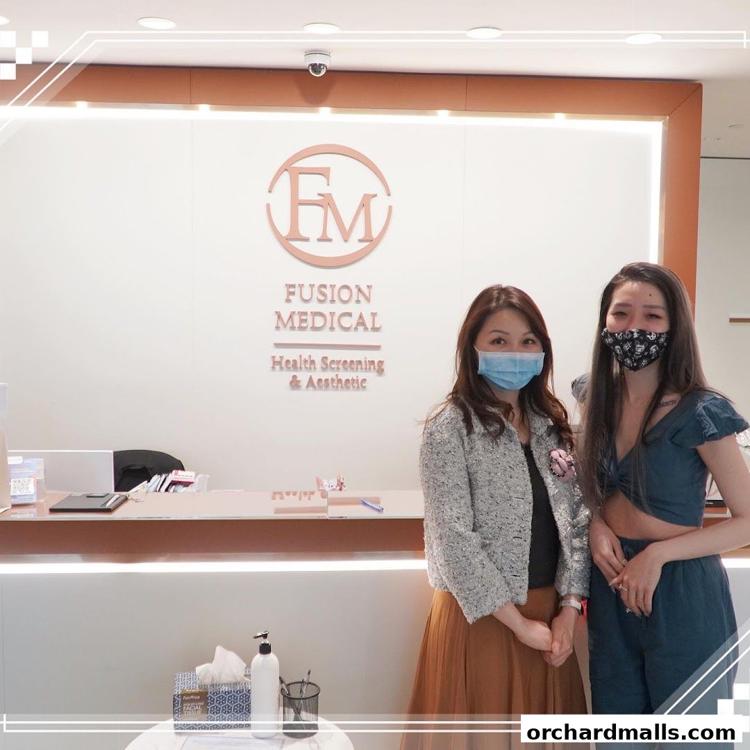 Two women stand in front of the Fusion Medical reception desk, which displays health screening and aesthetic services.