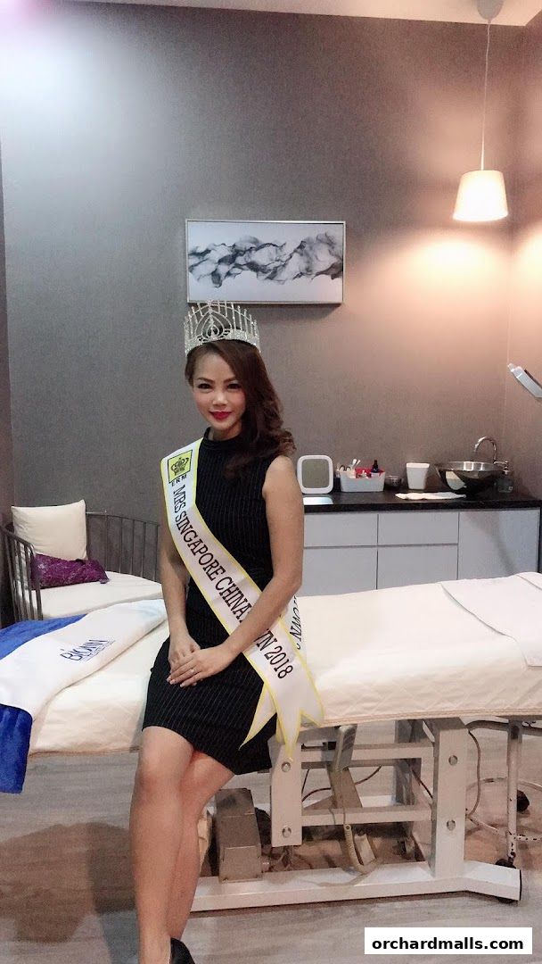 A woman wearing a crown and sash sits on a medical bed in a salon.