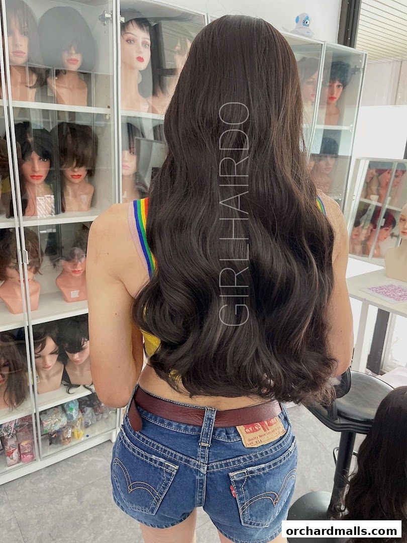 A person models a long, dark, wavy wig in a wig shop with many wigs displayed.