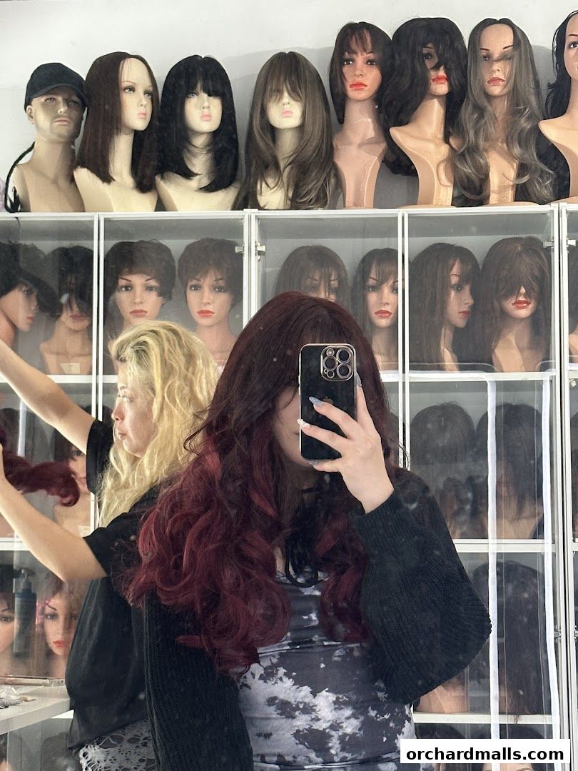 A person takes a selfie in a wig shop, surrounded by mannequins displaying various wigs.