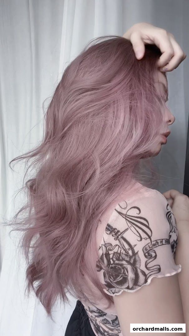 A person with long, wavy, pink-toned hair touching their head.