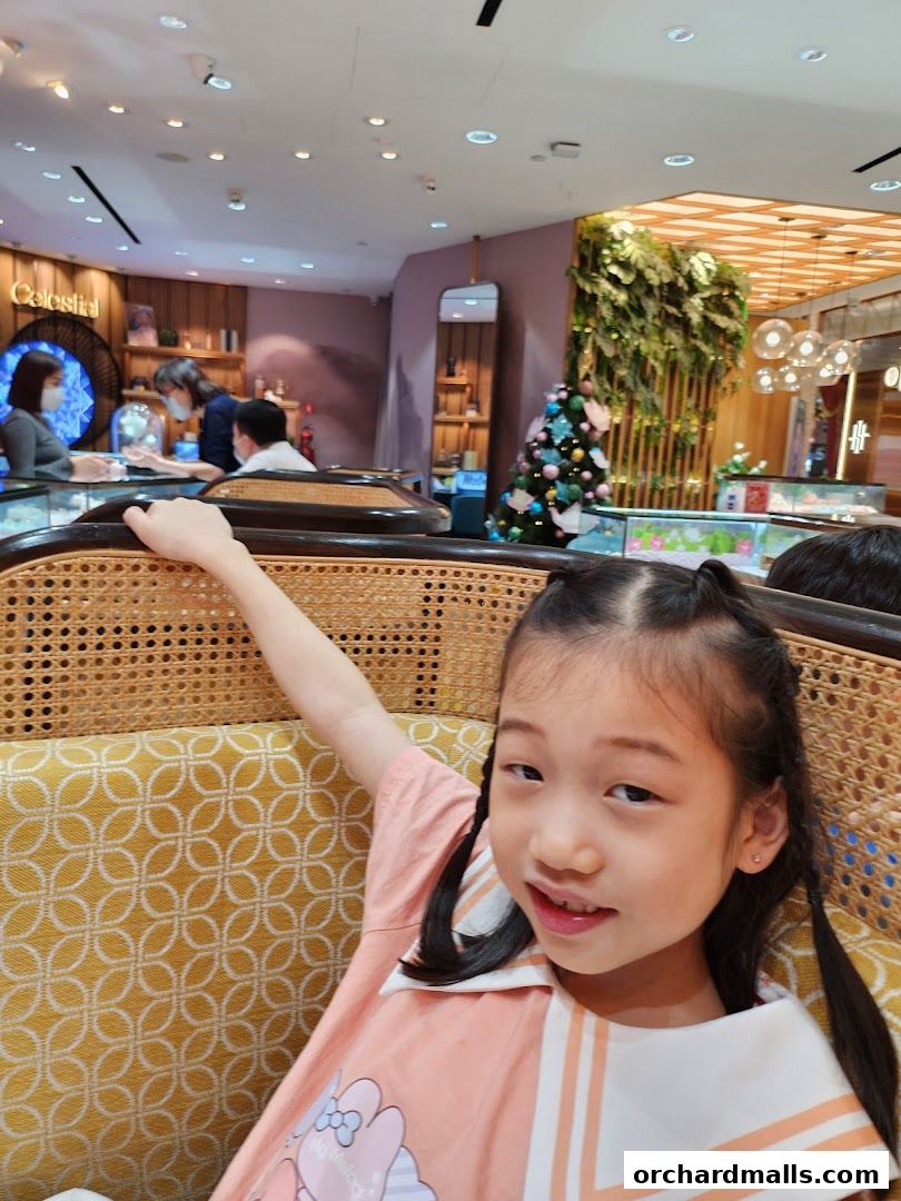 A young girl smiles while sitting in a jewelry store with displays and a Christmas tree in the background.