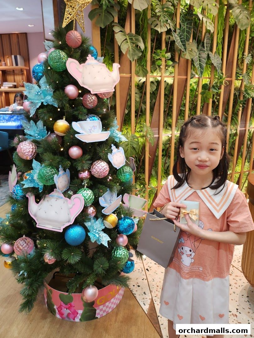 A young girl holds a gift and shopping bag next to a decorated Christmas tree.
