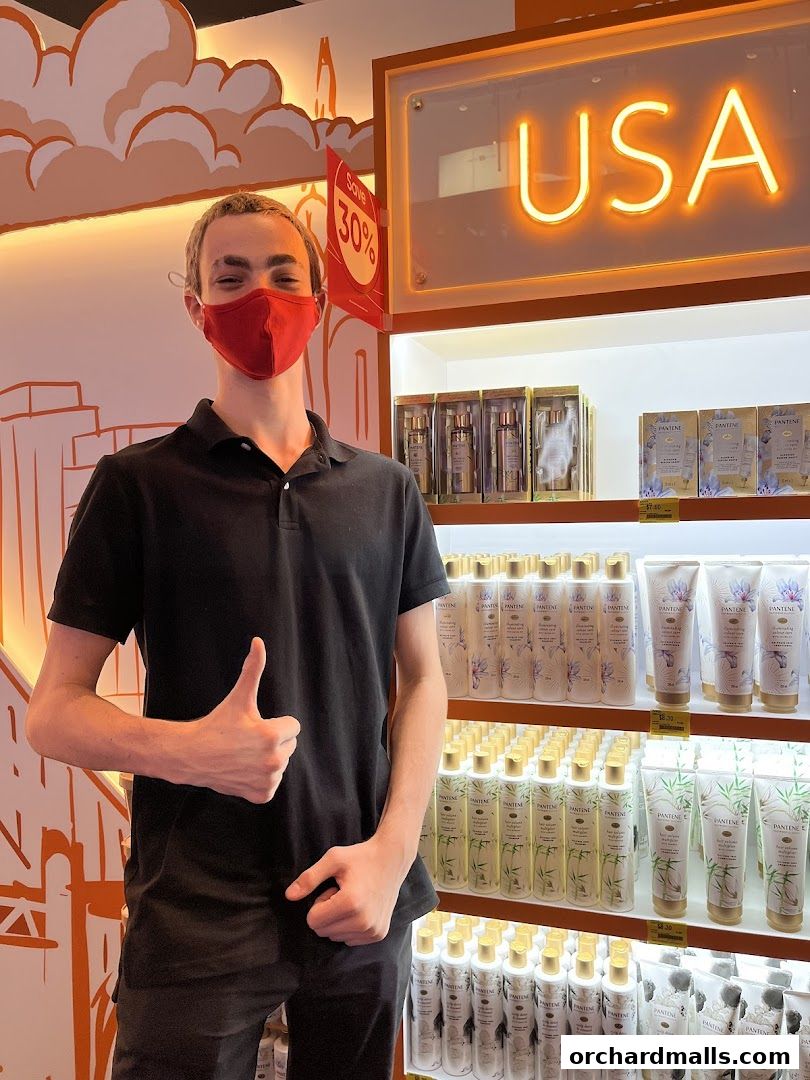 A person wearing a mask gives a thumbs up in front of shelves stocked with beauty products.