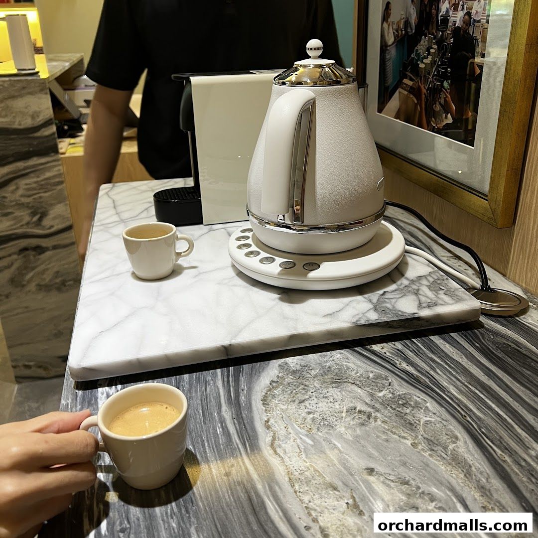 A hand holds a cup of coffee next to a kettle and coffee machine.