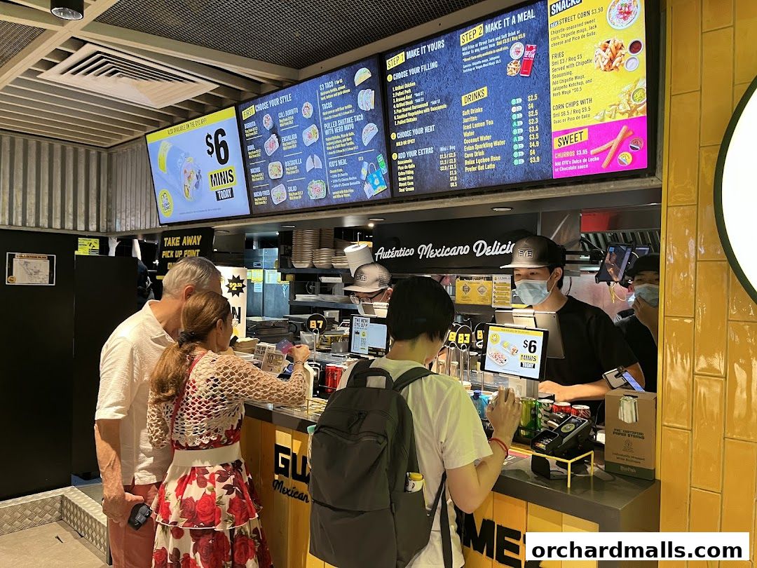Customers ordering at a Guzman y Gomez counter with a large digital menu board above.