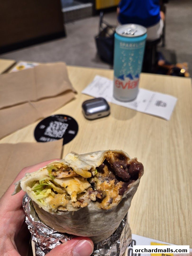 A hand holds a partially eaten burrito with chicken, beans, and lettuce, next to a can of Evian sparkling water.