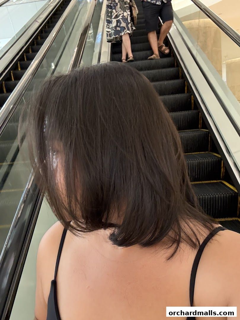 A person with dark hair stands in front of an escalator with other shoppers.