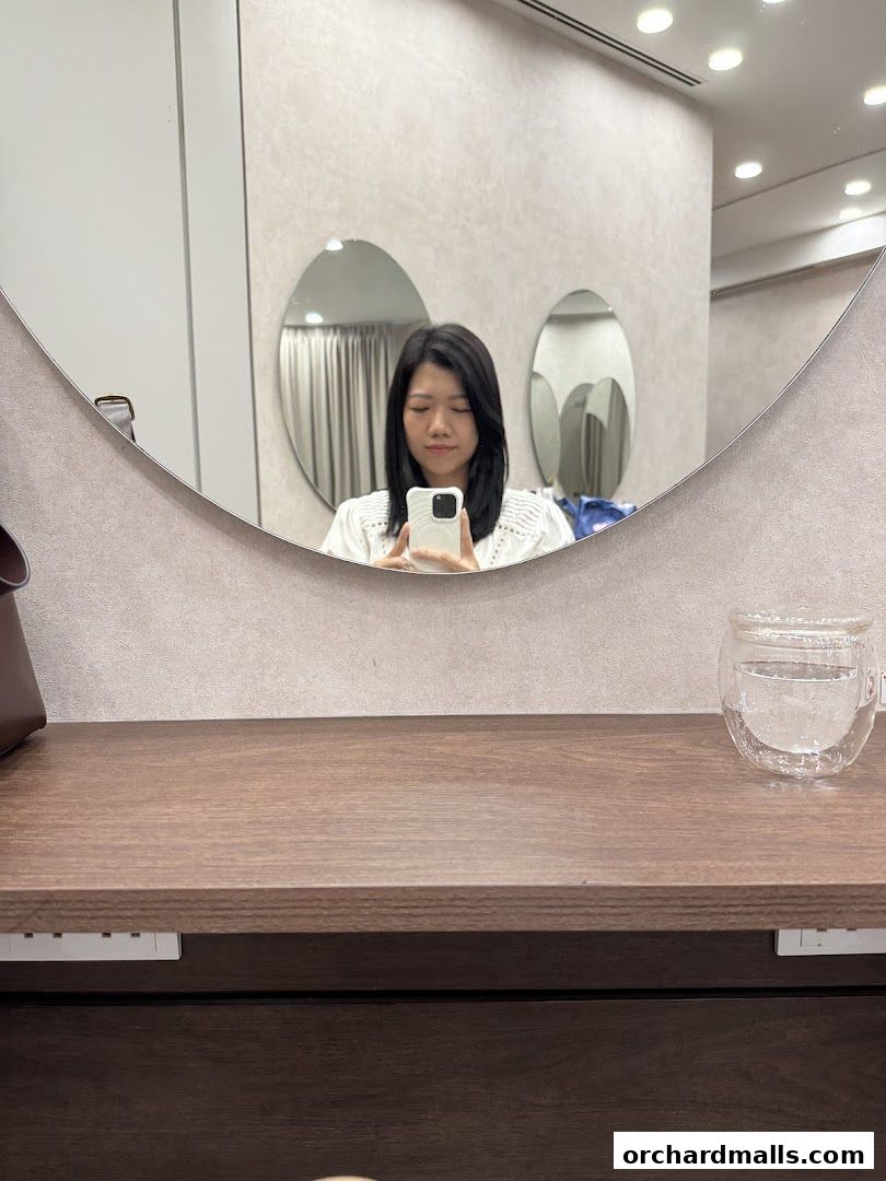 A woman takes a selfie in a mirror at a salon, with a glass of water on the counter.