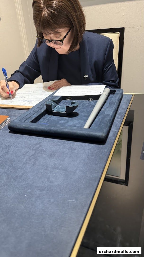 A woman in a navy blazer signs documents at a desk with a jewelry display.