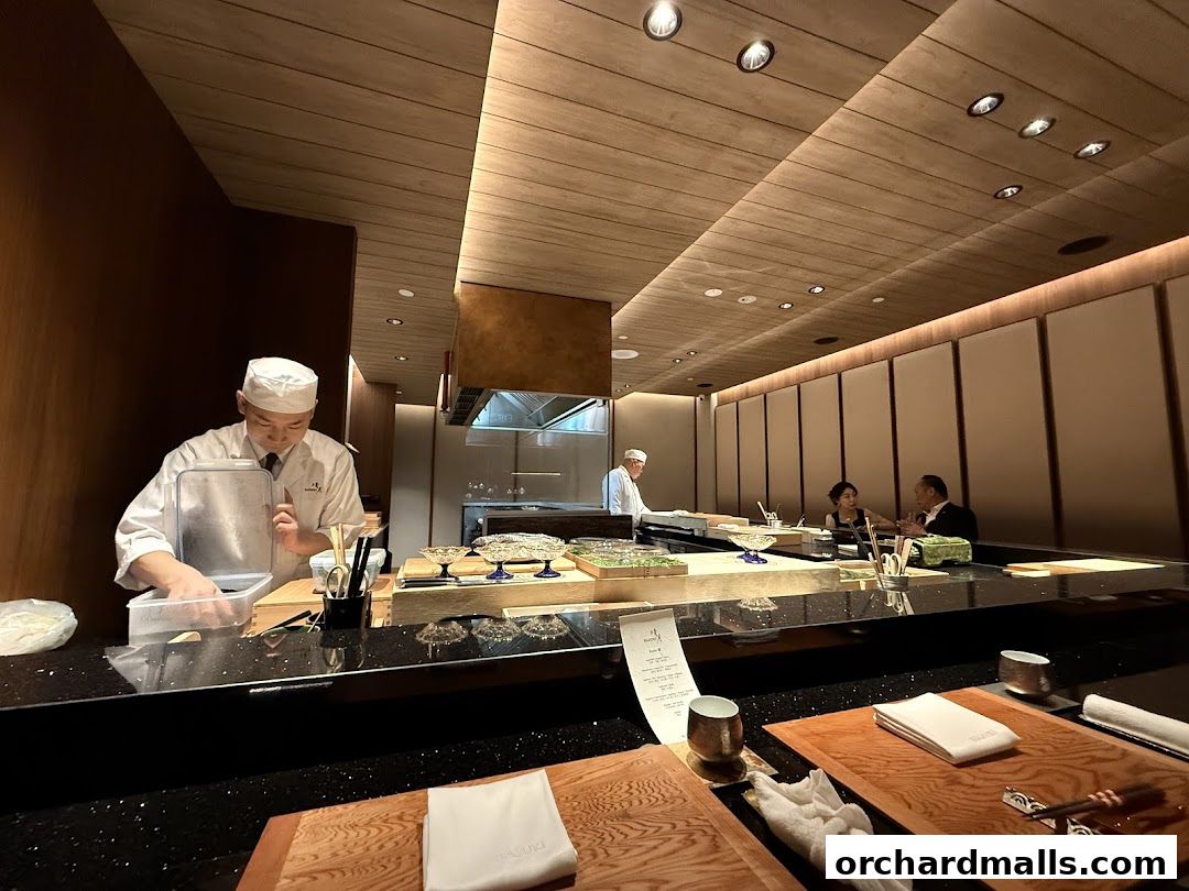 Chefs prepare food at a sushi bar while diners are seated at the counter.