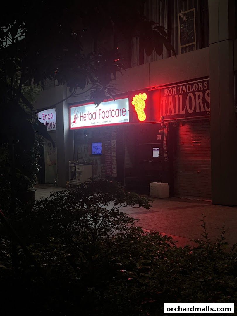 The illuminated storefront of Herbal Footcare and Ron Tailors at night.