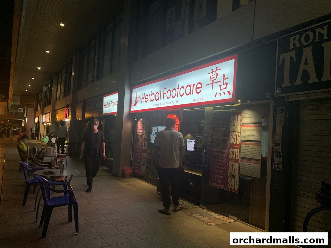 The storefront of Herbal Footcare, a health and beauty center, at night with prices displayed.