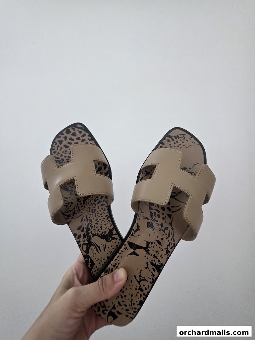 A hand holds a pair of beige Hermès sandals with a leopard print sole.