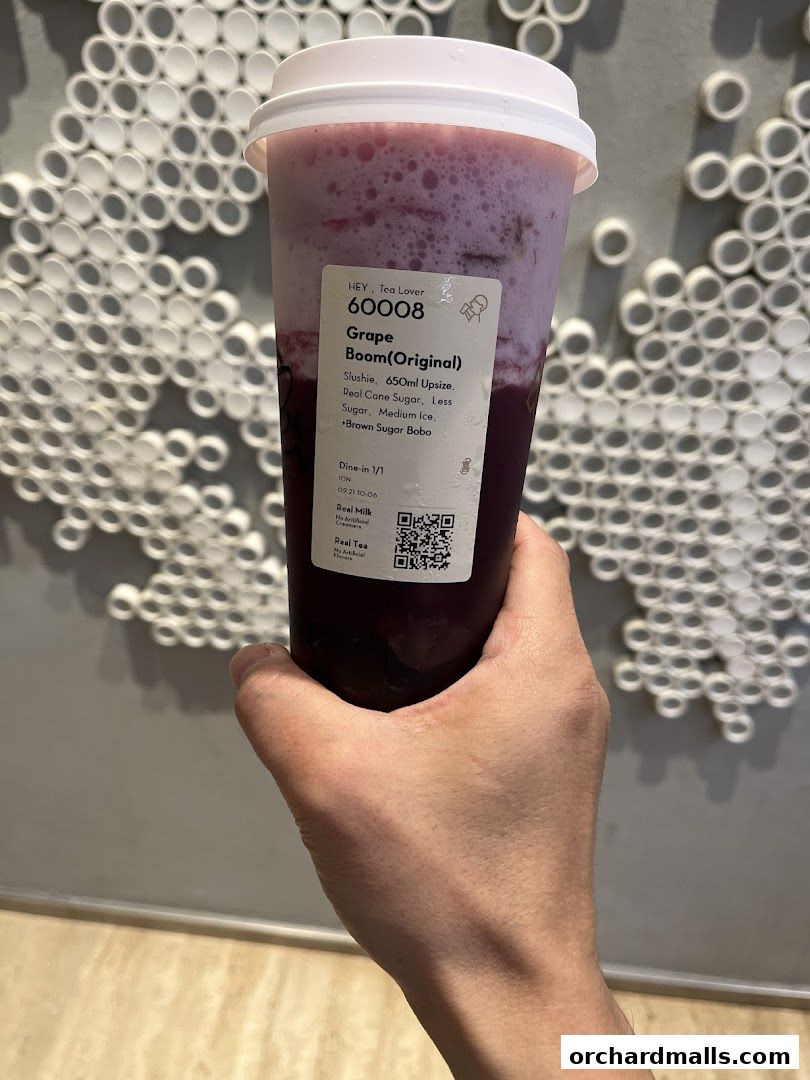 A hand holds a purple Grape Boom slushie from HEYTEA with a white lid.