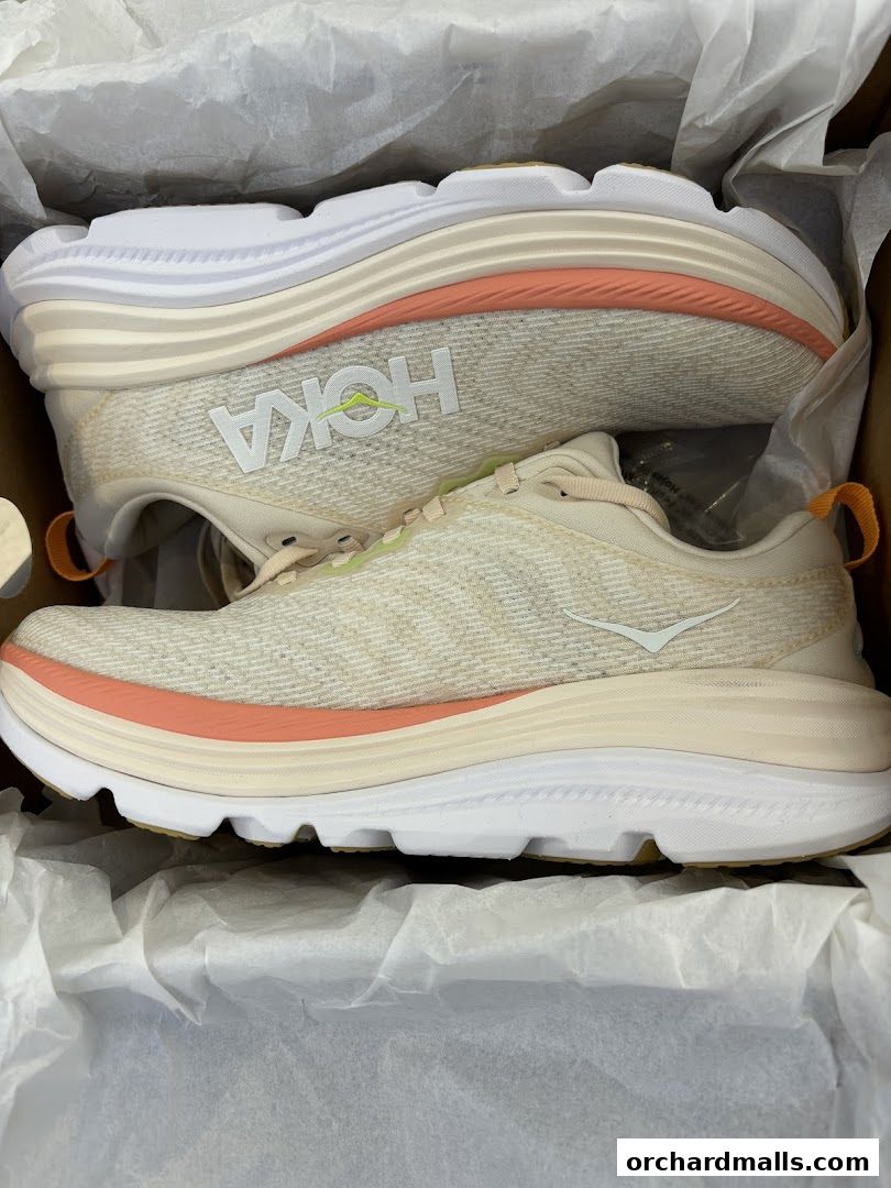 A pair of Hoka running shoes in a box, featuring a light beige color with coral accents.