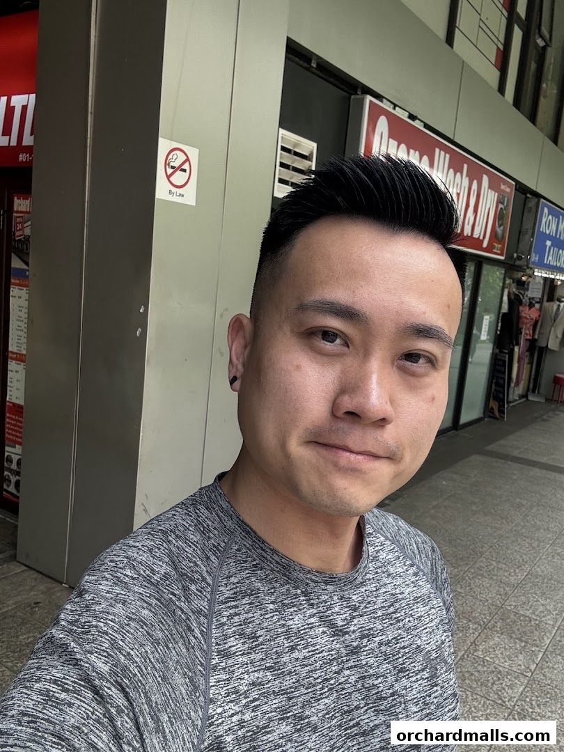 A man in a grey t-shirt poses for a selfie in front of a barbershop.