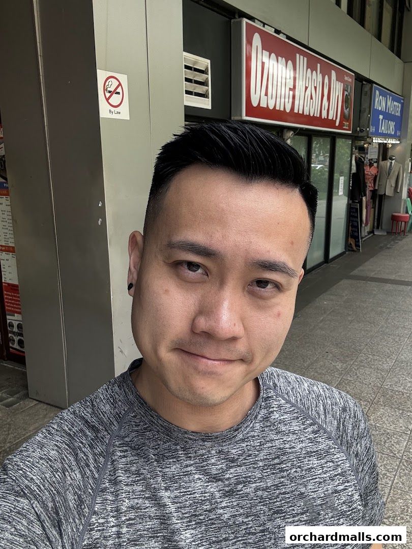 A man smiles at the camera in front of a barbershop and other retail stores.