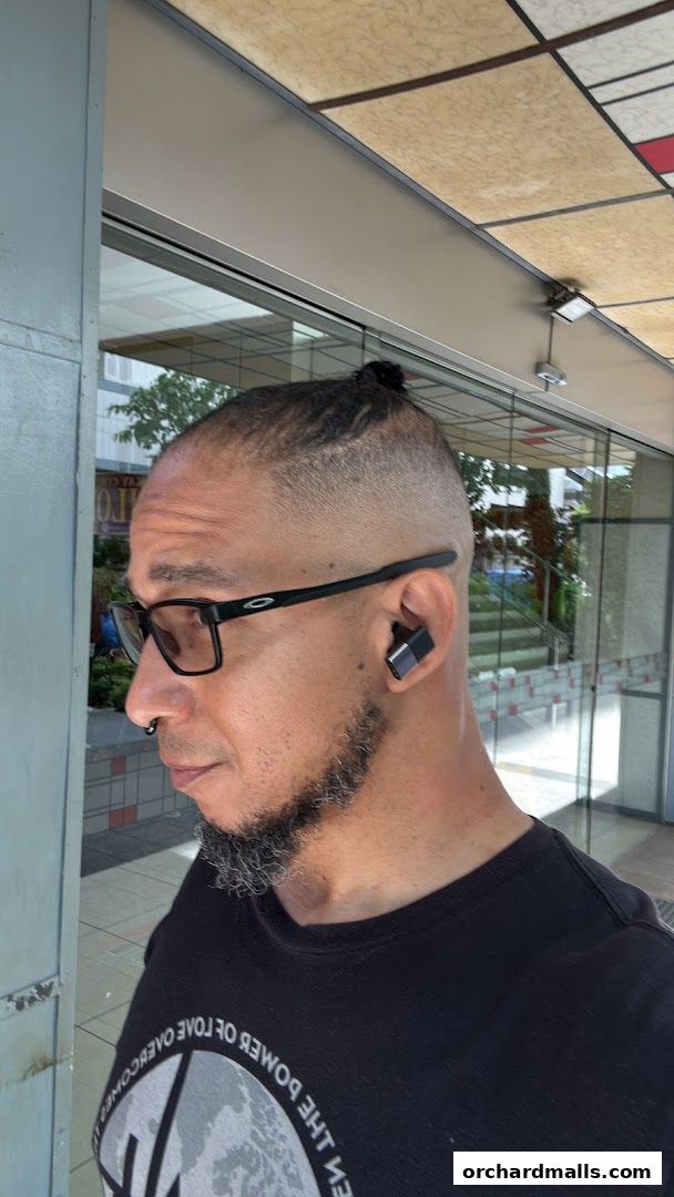 A man with a top knot hairstyle and glasses stands outside a shop.