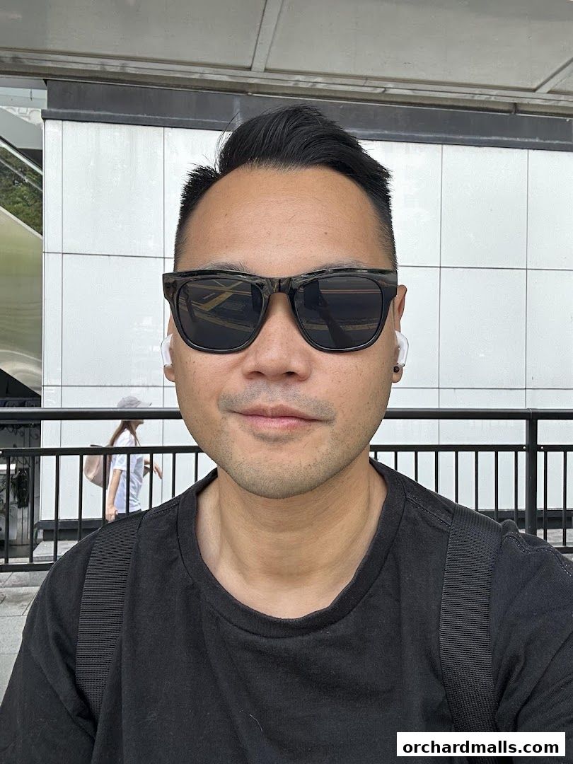 A man wearing sunglasses and a black t-shirt poses for a selfie outdoors.