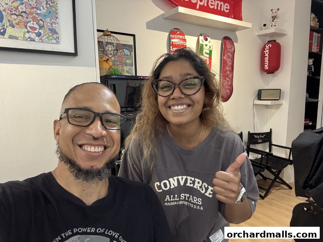 Two smiling people give a thumbs up in a barbershop with skateboards and Supreme merchandise on display.