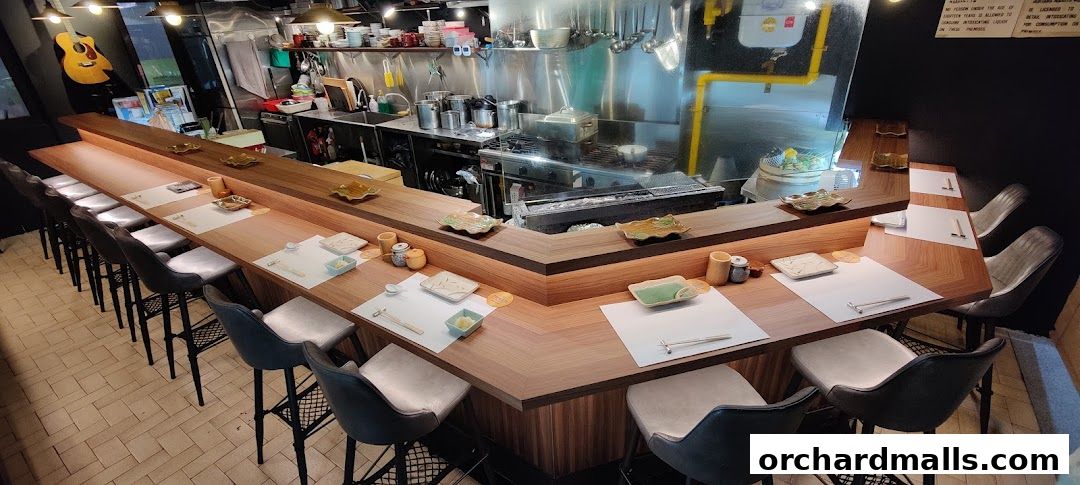 Interior of an izakaya with a long wooden counter, bar stools, and a kitchen in the background.