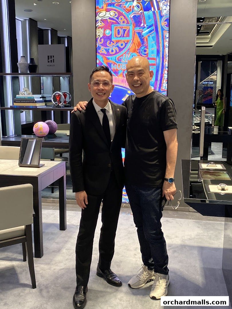 Two men smiling in a luxury watch boutique with a large digital display.
