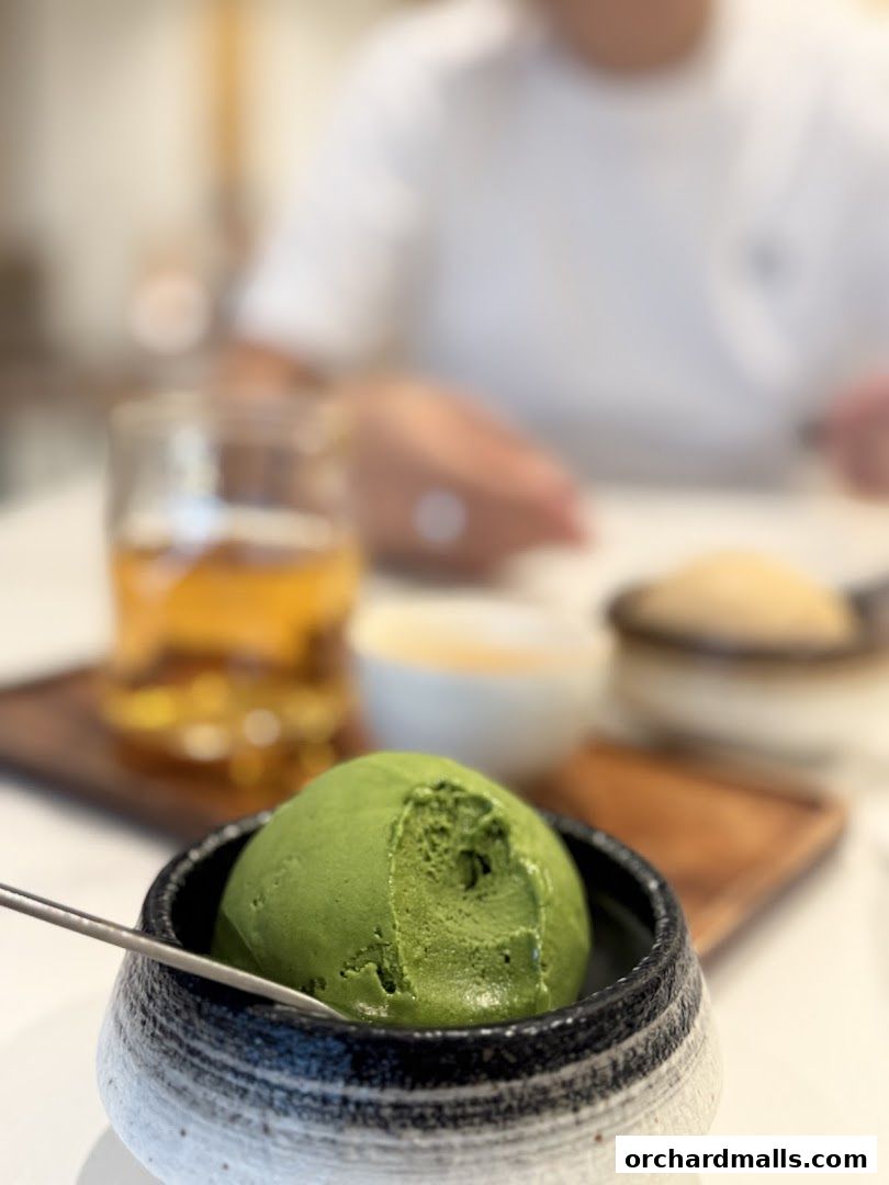 A scoop of matcha ice cream in a bowl with a drink and dessert in the background.