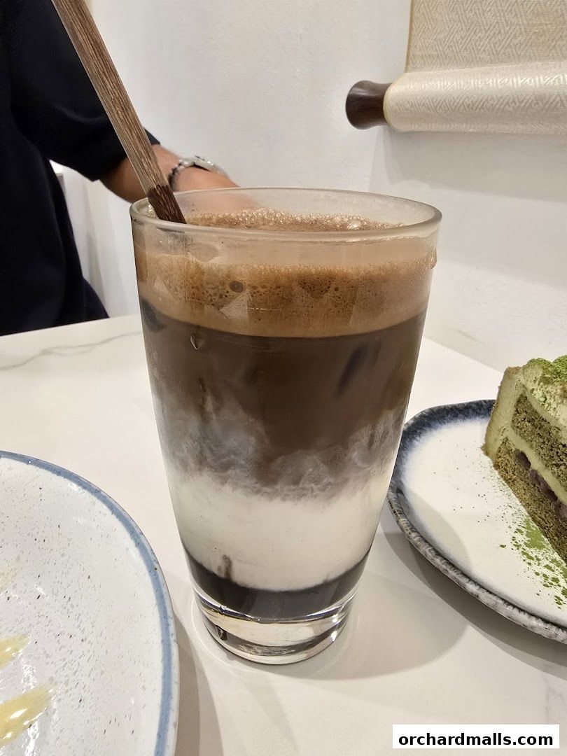 A layered iced coffee drink with a wooden stirrer and a slice of matcha cake.