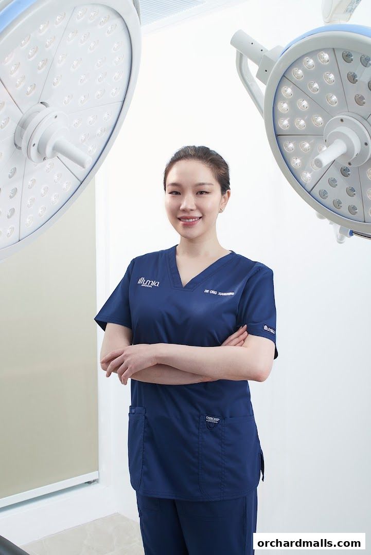 A smiling medical professional in scrubs stands with arms crossed under surgical lights.