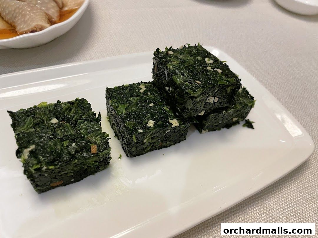 A white plate holds several square-shaped portions of finely chopped green vegetables with visible pieces of garlic.