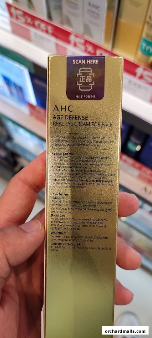 A hand holds a box of AHC Age Defense Real Eye Cream for Face.