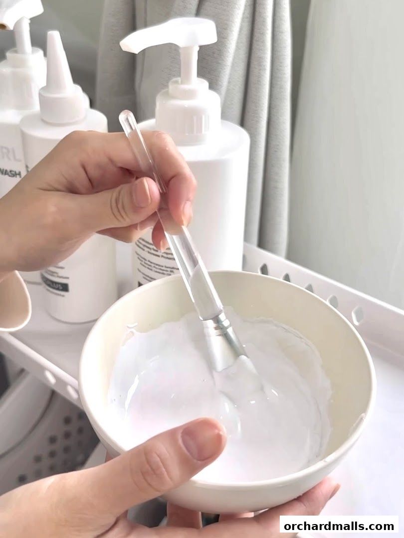 Hands mixing a white cream in a bowl with a brush, surrounded by beauty products.