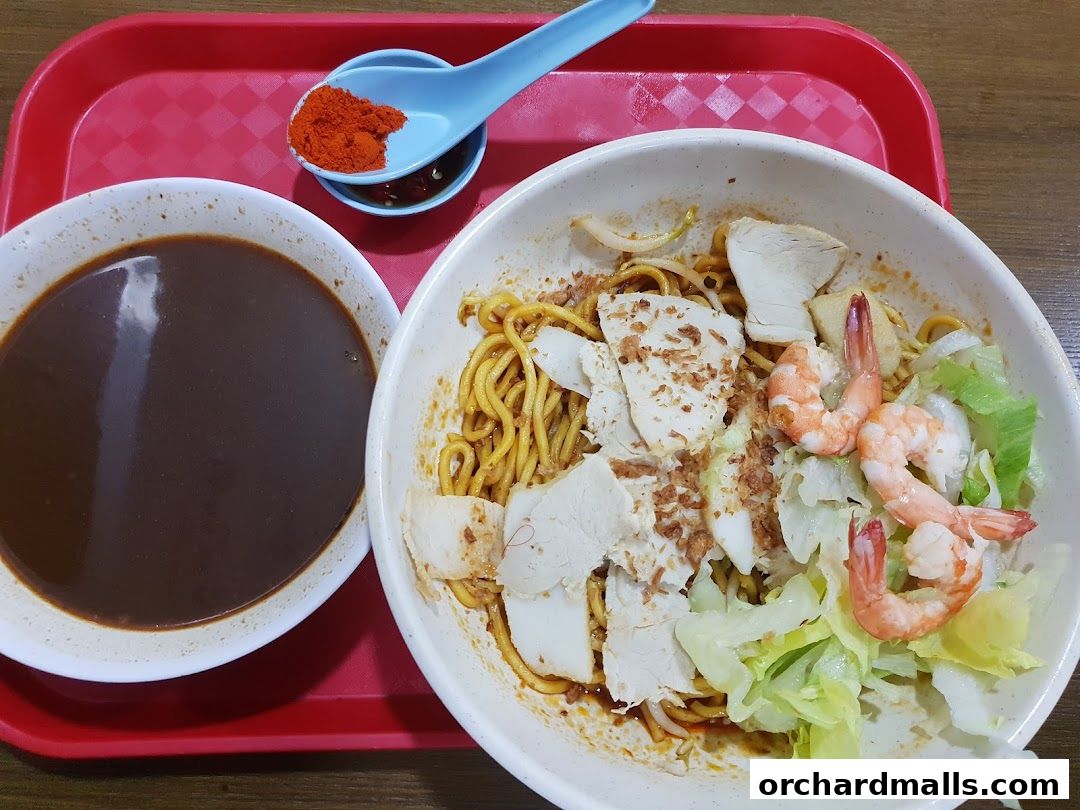 A bowl of noodles with chicken, shrimp, and vegetables, served with a side of dark sauce and chili.
