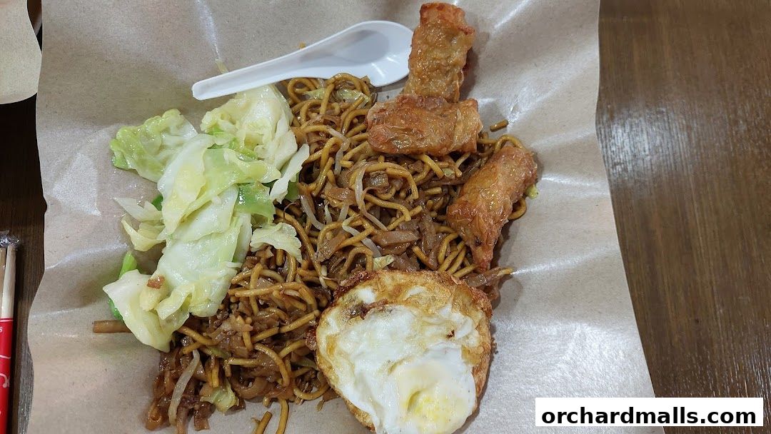 A plate of noodles with fried egg, cabbage, and fried spring rolls served on brown paper.