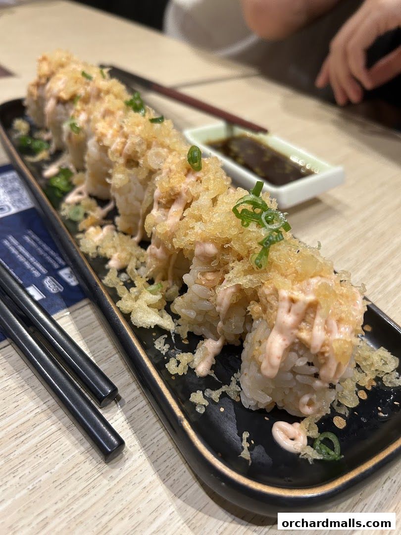 A close-up of a sushi roll with crispy toppings and sauce, served with soy sauce and chopsticks.