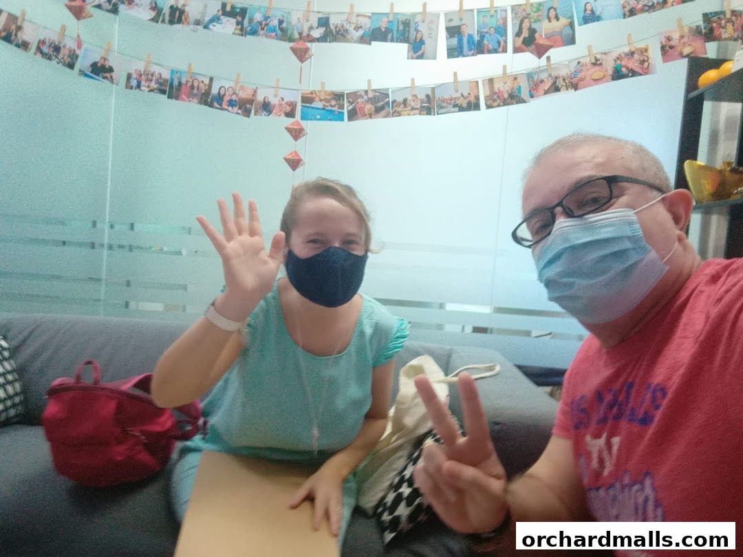 Two people wearing masks wave hello in a room decorated with photos.