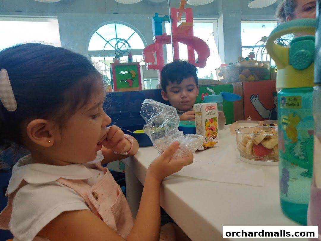 Children eating snacks at a table with toys and drinks in the background.