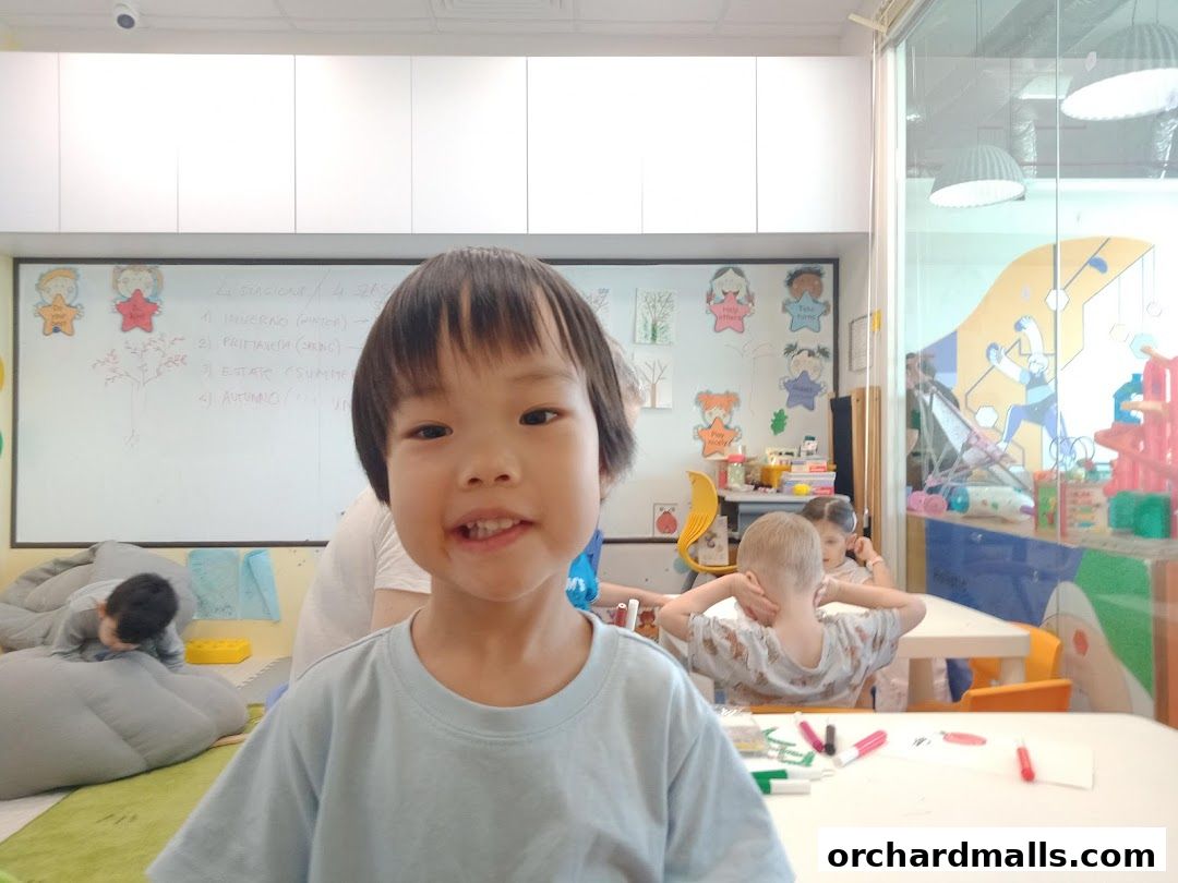 A young child smiles at the camera in a classroom with a whiteboard and other children in the background.