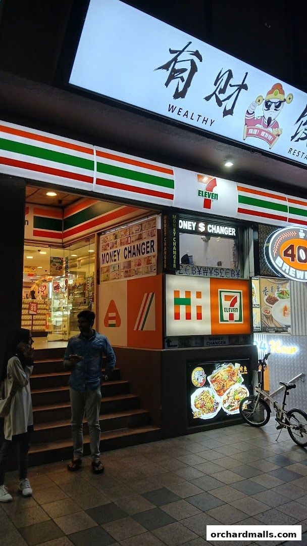 A 7-Eleven convenience store with a money changer service and food advertisements.