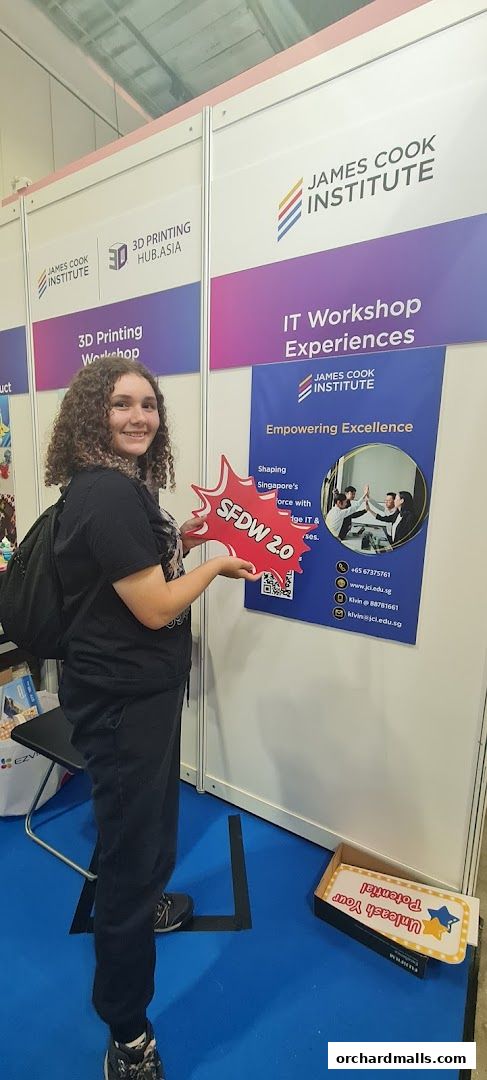 A young woman holds a 'SFDW 2.0' sign in front of a James Cook Institute booth.
