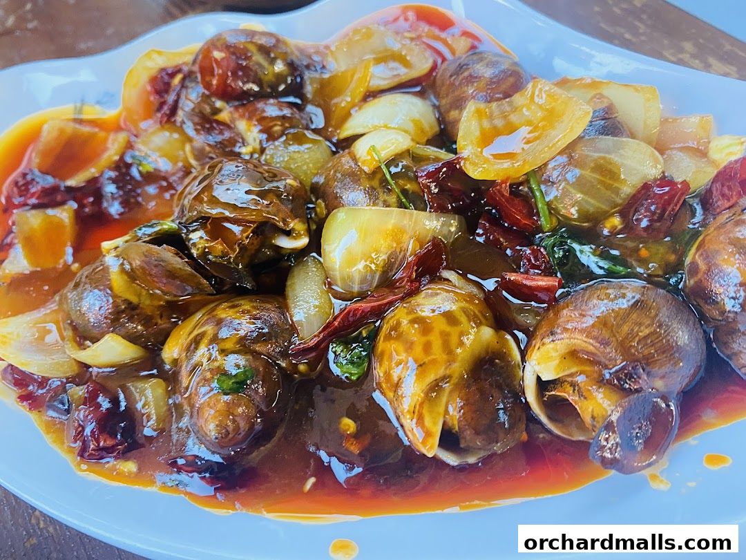 A close-up of a plate of escargots in a rich, savory sauce with onions and chili peppers.