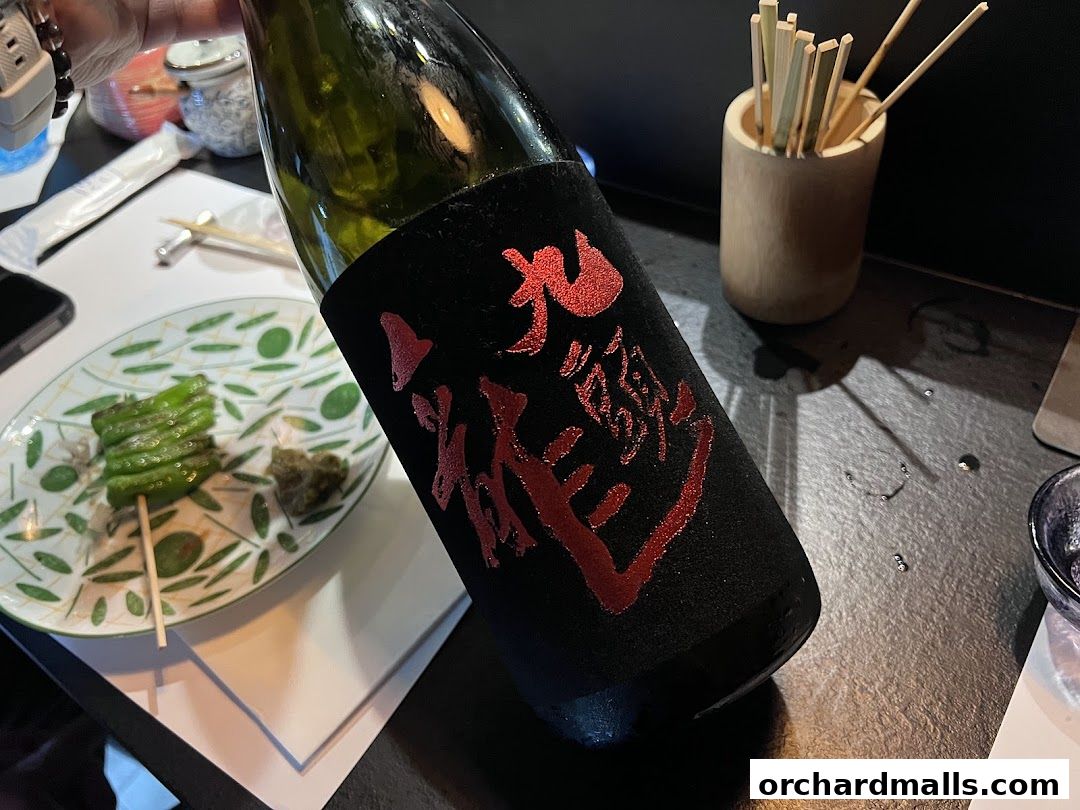 A bottle of sake with red Japanese calligraphy sits next to a skewer of grilled green peppers.