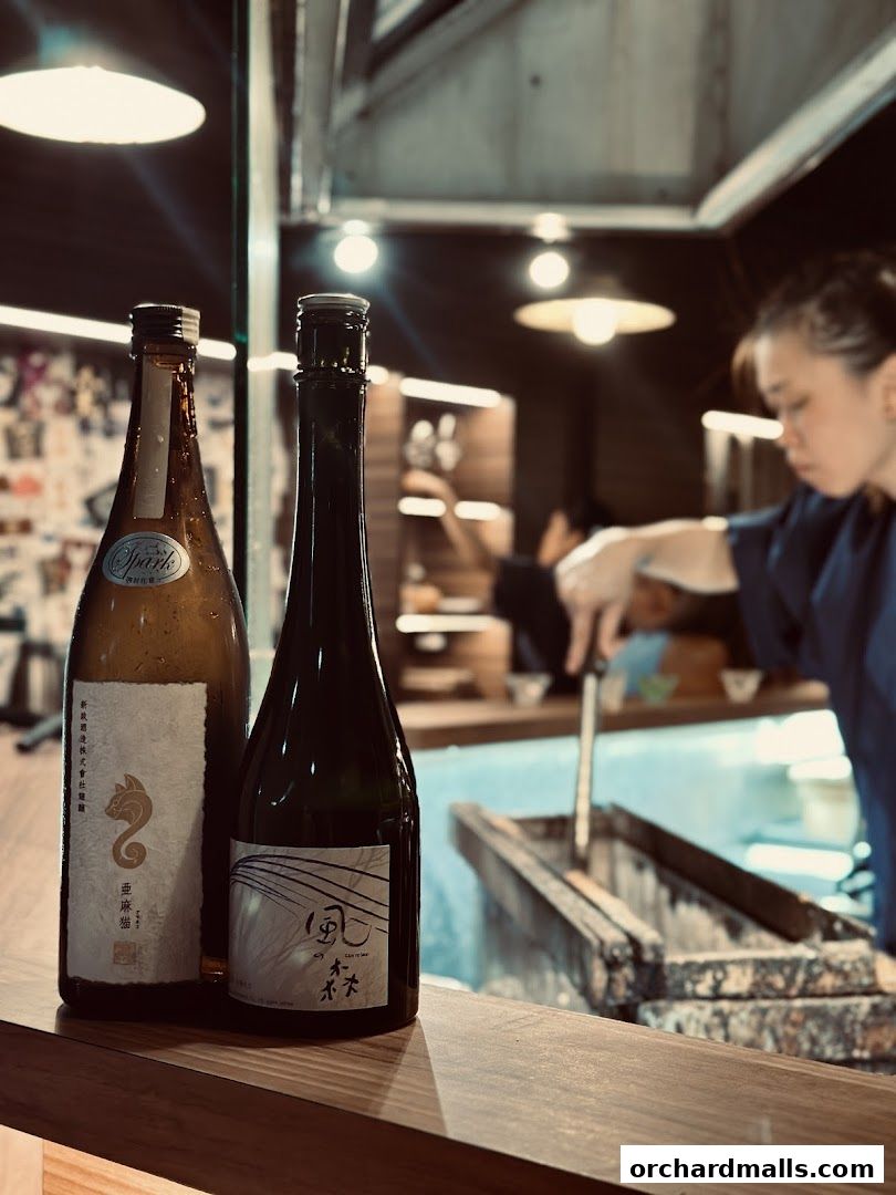 Two bottles of sake are displayed on a wooden counter with a chef preparing food in the background.