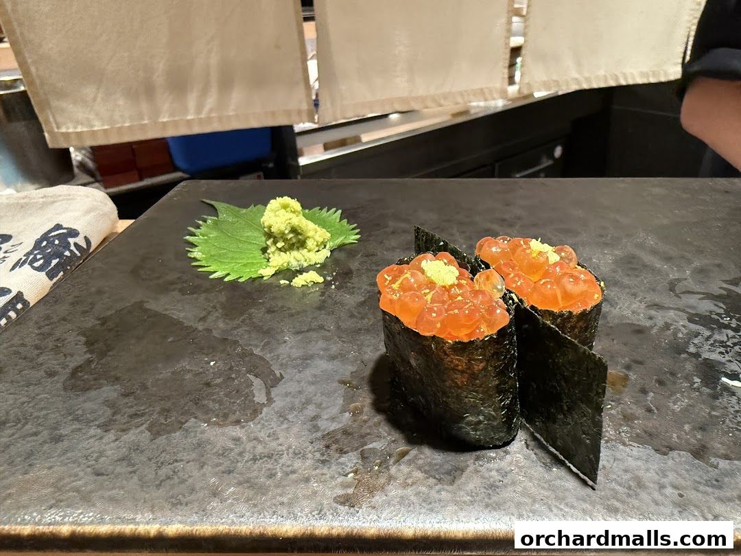 Two pieces of ikura sushi with wasabi and a perilla leaf on a dark surface.