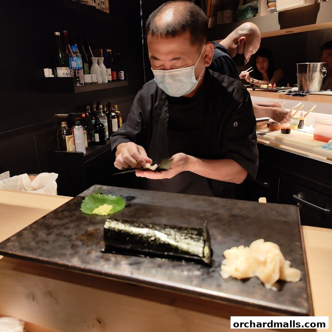 A sushi chef prepares a roll while other diners are visible in the background.