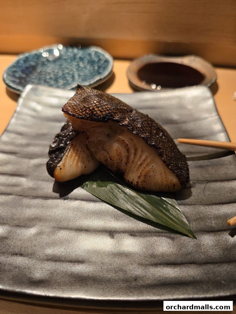 Two pieces of grilled fish with crispy skin served on a dark plate with a leaf garnish.