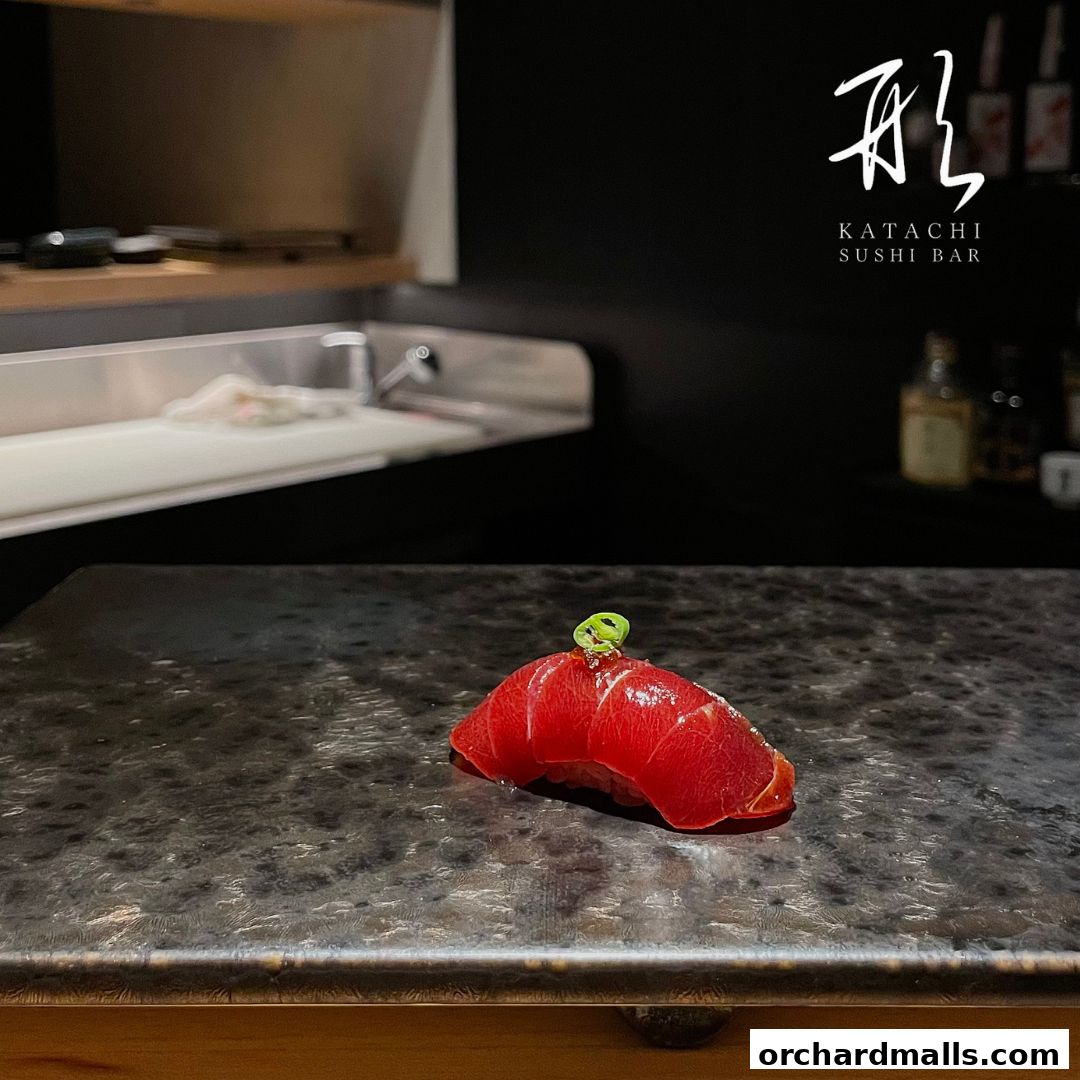 A single piece of tuna sushi with a garnish sits on a dark textured plate.