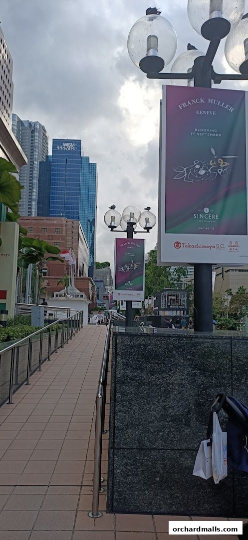 A walkway leads towards a shopping mall with tall buildings and advertising banners.
